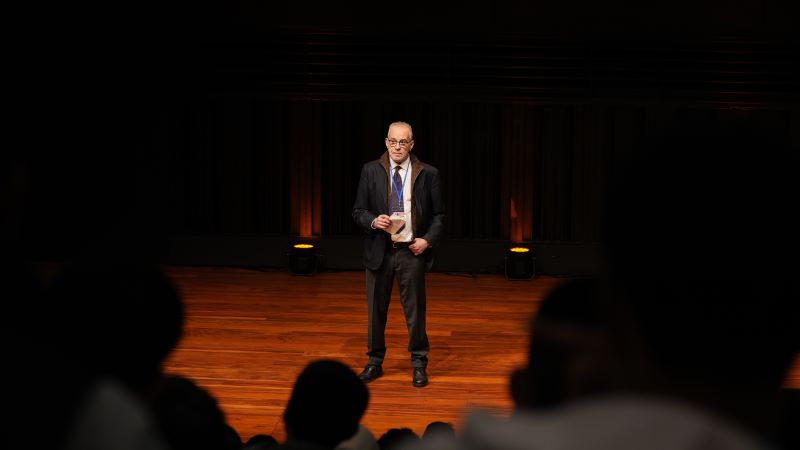 Persona en escenario de auditorio con acreditaci&oacute;n, hablando al p&uacute;blico con iluminaci&oacute;n c&aacute;lida y espectadores en primer plano desenfocados