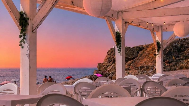 Sobrevent, un restaurante que da vida a la playa Interior de chiringuito junto al mar al atardecer, con mesas y sillas blancas bajo una pérgola de madera, vistas al Mediterráneo y acantilado rocoso.