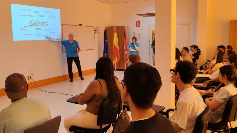 Daniel Jiménez, exempleado de CaixaBank que ofrece formación a personas en situación de vulnerabilidad y jóvenes estudiantes Voluntario impartiendo una charla ante un grupo en un aula, señalando una presentación proyectada sobre gastos; asistentes sentados escuchan la exposición