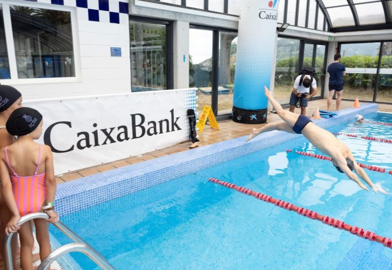 Actividad de nataci&oacute;n en una piscina cubierta; &Iacute;&ntilde;igo Llopis se lanza al agua mientras varios ni&ntilde;os observan junto a un panel de CaixaBank.