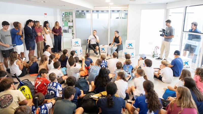 Charla educativa ante un numeroso grupo de ni&ntilde;os sentados en el suelo, con dos ponentes al frente y equipos de grabaci&oacute;n en una sala de CaixaBank.