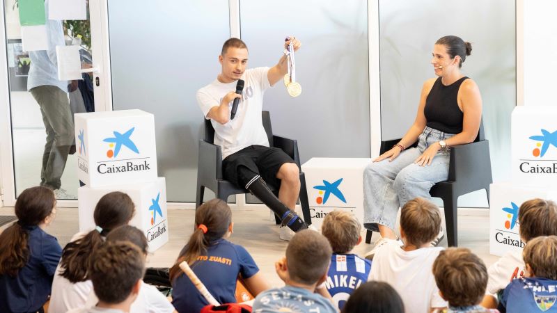 &Iacute;&ntilde;igo Llopis muestra unas medallas y habla con micr&oacute;fono ante un grupo de ni&ntilde;os sentados, en una actividad educativa con carteles de CaixaBank.