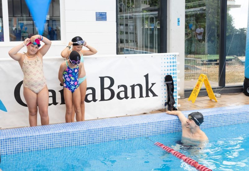 Actividad de nataci&oacute;n infantil en piscina cubierta; dos ni&ntilde;as con gorro esperan junto al borde mientras &Iacute;&ntilde;igo Llopis permanece en el agua, con se&ntilde;alizaci&oacute;n de CaixaBank al fondo